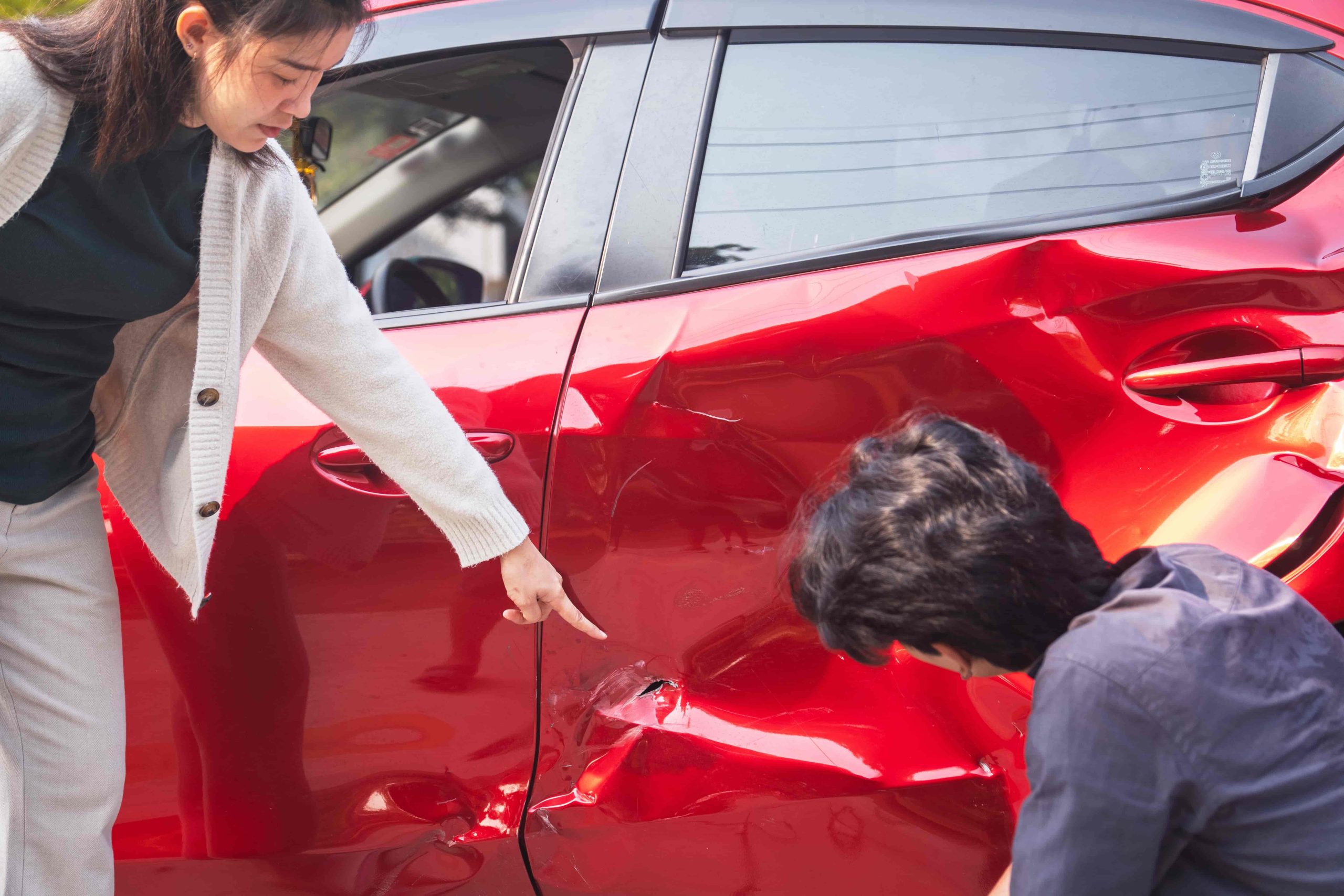 Woman pointing to car damage after accident while man inspects dent on red vehicle.